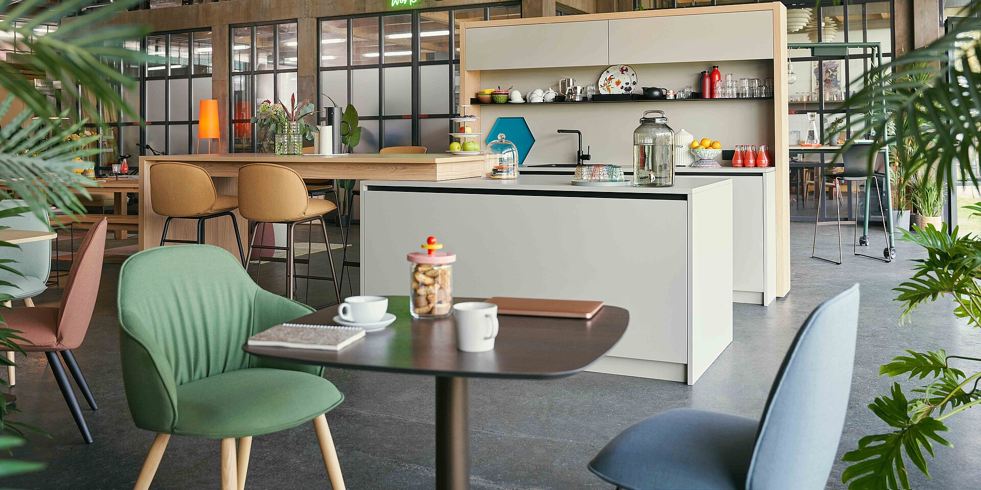 A modern communal space with a small kitchen, a table with green and pink chairs in the foreground, and additional seating areas in the background. Plants and warm lighting create a welcoming atmosphere.