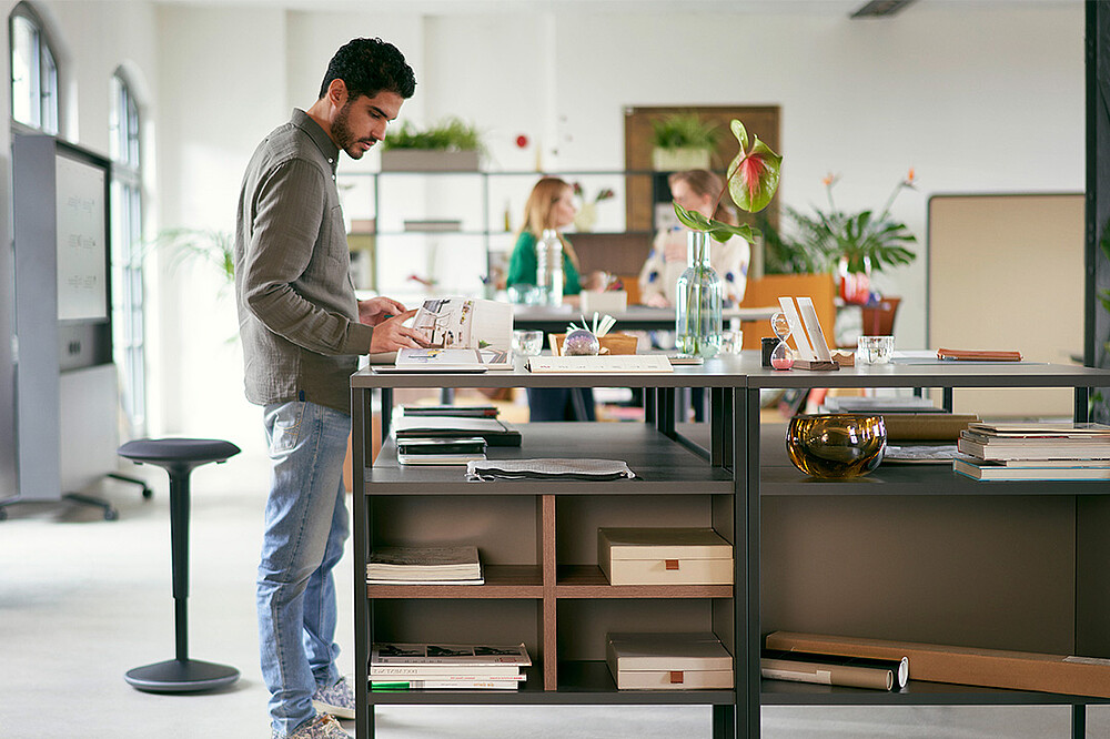 Ein Mann steht an einem se:matrix Regalsystem von Sedus und liest vertieft in einer Zeitschrift. Der Raum ist hell und modern gestaltet, durchflutet von natürlichem Licht. Im Hintergrund sind unscharf zwei Kolleginnen zu sehen, die sich unterhalten, was dem Raum eine lebendige und kommunikative Atmosphäre verleiht. Hinter dem Mann steht eine se:fit von Sedus, die flexible Arbeitsweisen unterstützt. Diese Szene verdeutlicht das Konzept der hybriden Konfigurationen, in denen flexible Arbeitsumgebungen geschaffen werden, um Produktivität und Wohlbefinden der Mitarbeiter zu fördern, inspiriert von Cafés, städtischen Räumen und klassischen Büros.