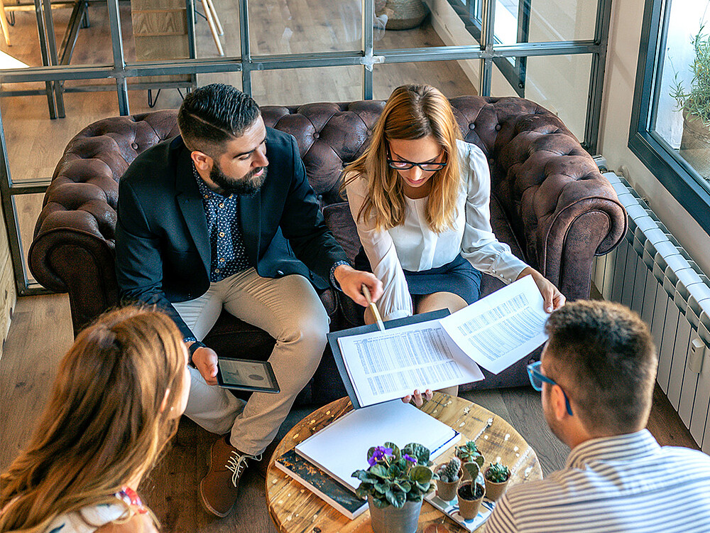 Ansicht von oben auf ein Team aus vier Personen, die sich auf einem Ledersofa über Dokumente unterhalten. Ein Mann hält ein Tablet in der Hand und deutet mit einem Stift auf ein Dokument. Diese Szene veranschaulicht das Konzept der hybriden Zeiten, in dem Arbeit und Privatleben in einem ökosystemischen Rahmen vereint werden. Das ideale Büro wird als kommunikativer und einladender Knotenpunkt dargestellt, eingebettet in ein flexibles Netzwerk aus verschiedenen Arbeitsräumen, einschließlich Dritträumen und Home-Office, das eine ganzheitliche und umweltfreundliche Lebensweise fördert.