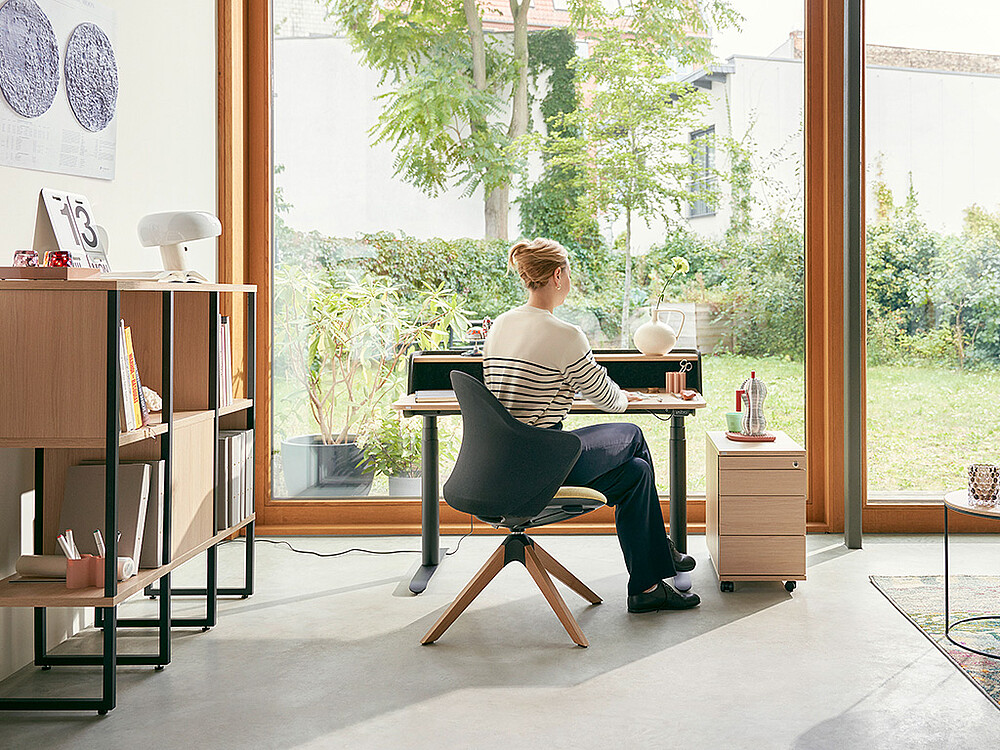Eine Frau arbeitet konzentriert in ihrem Homeoffice, das mit einem se:desk home-Schreibtisch und einem se:flair ausgestattet ist. Durch ein großes Fenster vor ihr strömt natürliches Licht herein und bietet einen beruhigenden Blick auf den Garten. Links neben dem Arbeitsplatz sorgt das se:matrix für Ordnung und Struktur, während rechts ein schmaler Container zusätzlichen Stauraum bietet. Dieses Homeoffice spiegelt die moderne Anpassung an die neue Arbeitswelt wider, in der Flexibilität und Gastfreundschaft im Arbeitsumfeld zentrale Rollen spielen.