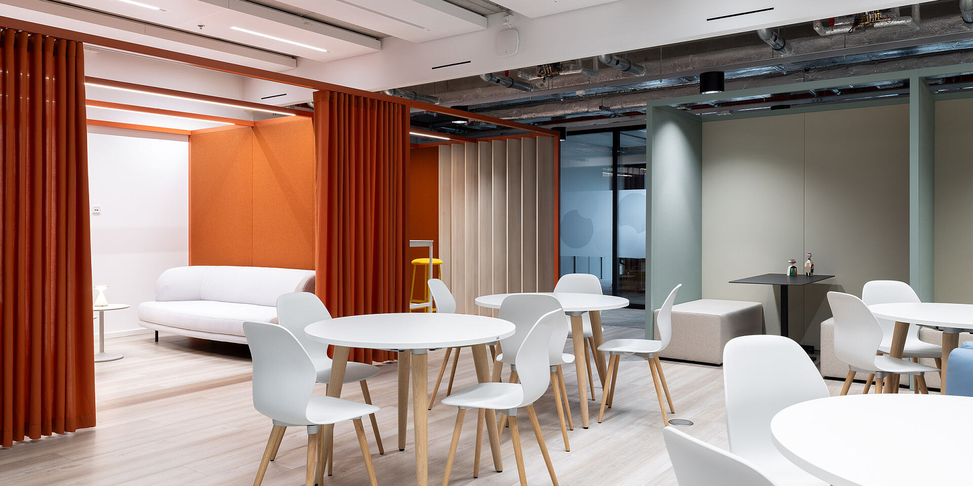 Moderner Bistrobereich bei Veolia mit weißen se:spot Designstühlen und se:assist Tischen, orangefarbene Wand und weiße Couch im Hintergrund